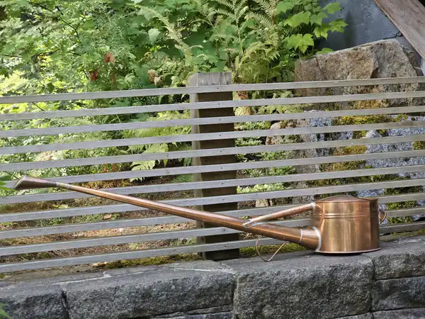 A copper watering can with a long spout resting on a stone wall in front of a slatted fence