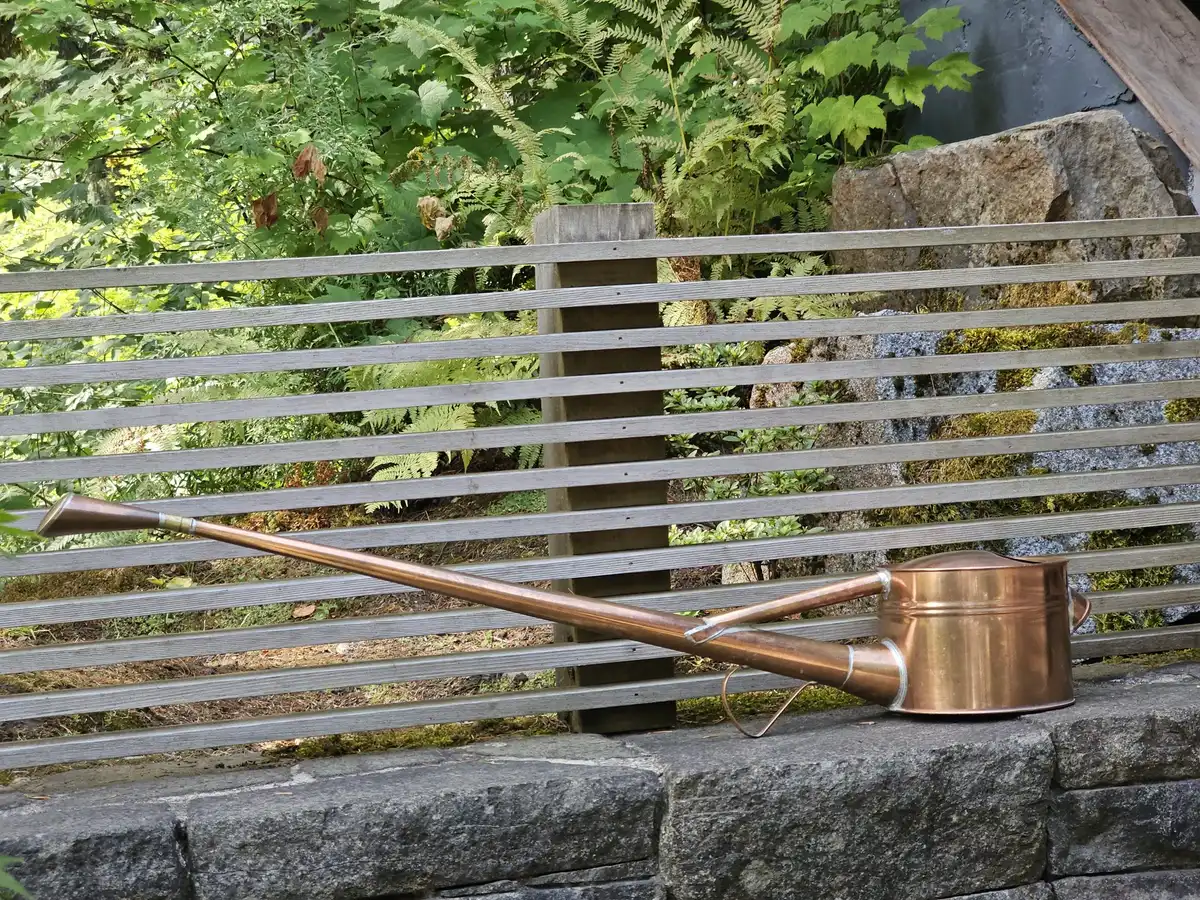A copper watering can with a long spout resting on a stone wall in front of a slatted fence